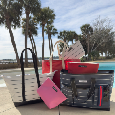 Bags displayed by a swimming pool, featuring different designs and colors, with water glistening nearby. Charleston Wrap fundraising, tote bag fundraiser